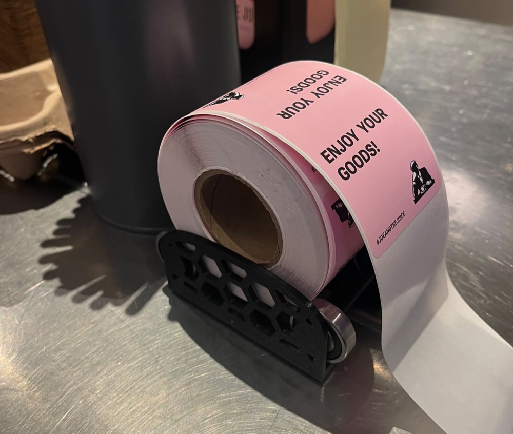 A 3D-printed black roller mount with a hexagonal lattice frame, holding a pink label roll on a bearing